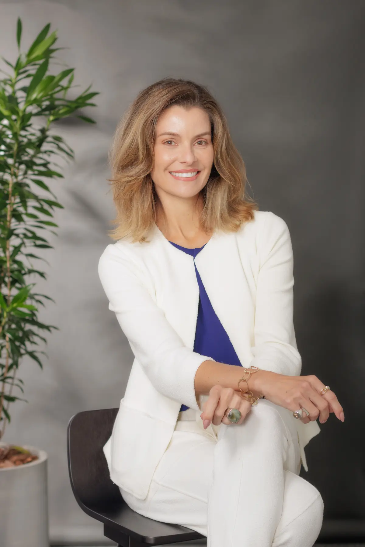 Executiva da Origem Energia em retrato institucional com traje formal, sentada em ambiente profissional com planta decorativa