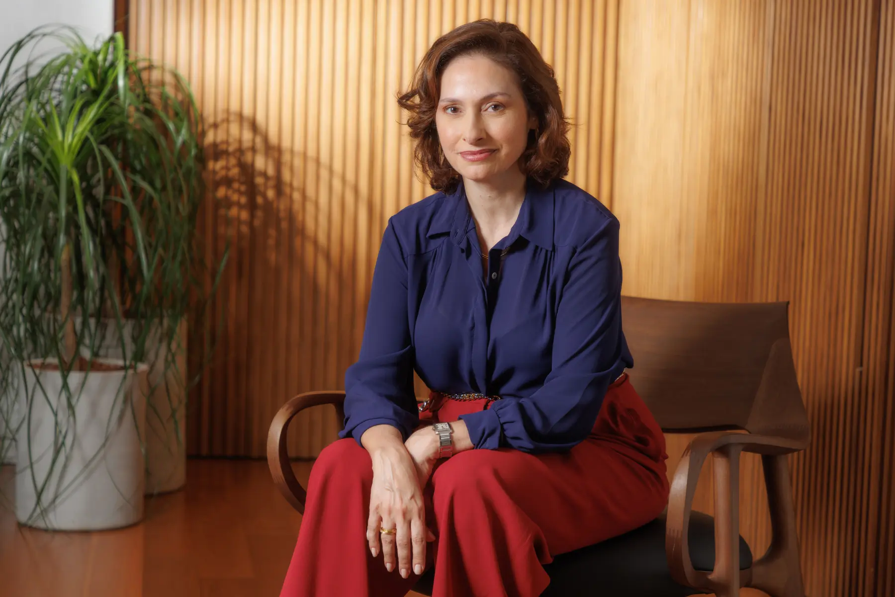 Executiva da Origem Energia em retrato institucional com traje formal, sentada em cadeira de madeira diante de parede com ripado e planta decorativa.