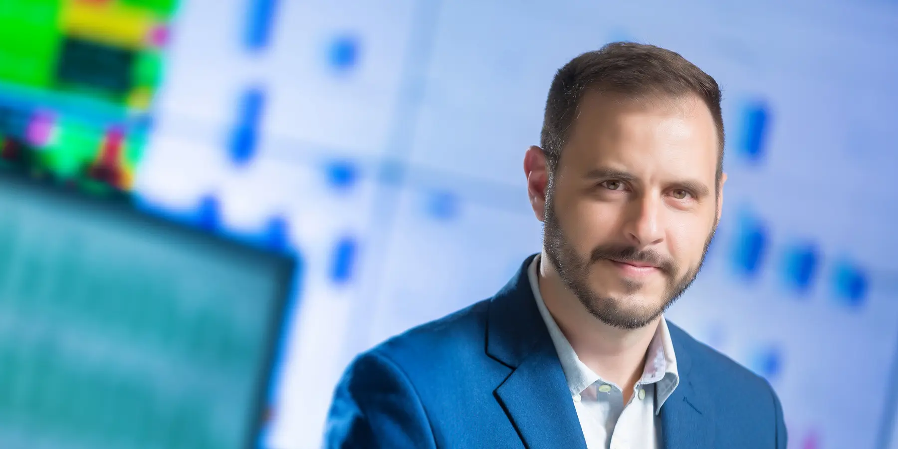 Retrato corporativo de um homem usando blazer azul em frente a tela com gráficos desfocados.