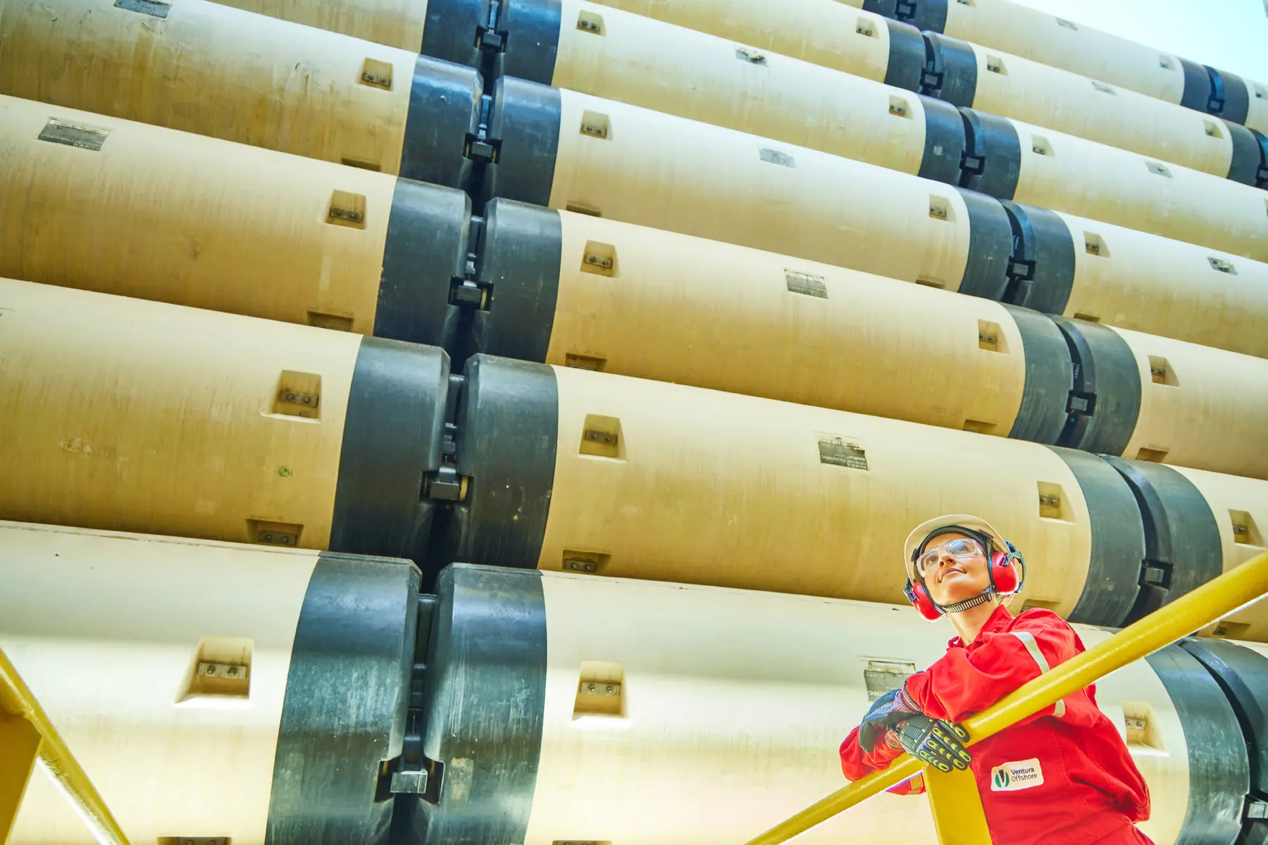Mulher offshore com EPIs diante de pilha de risers em estrutura industrial