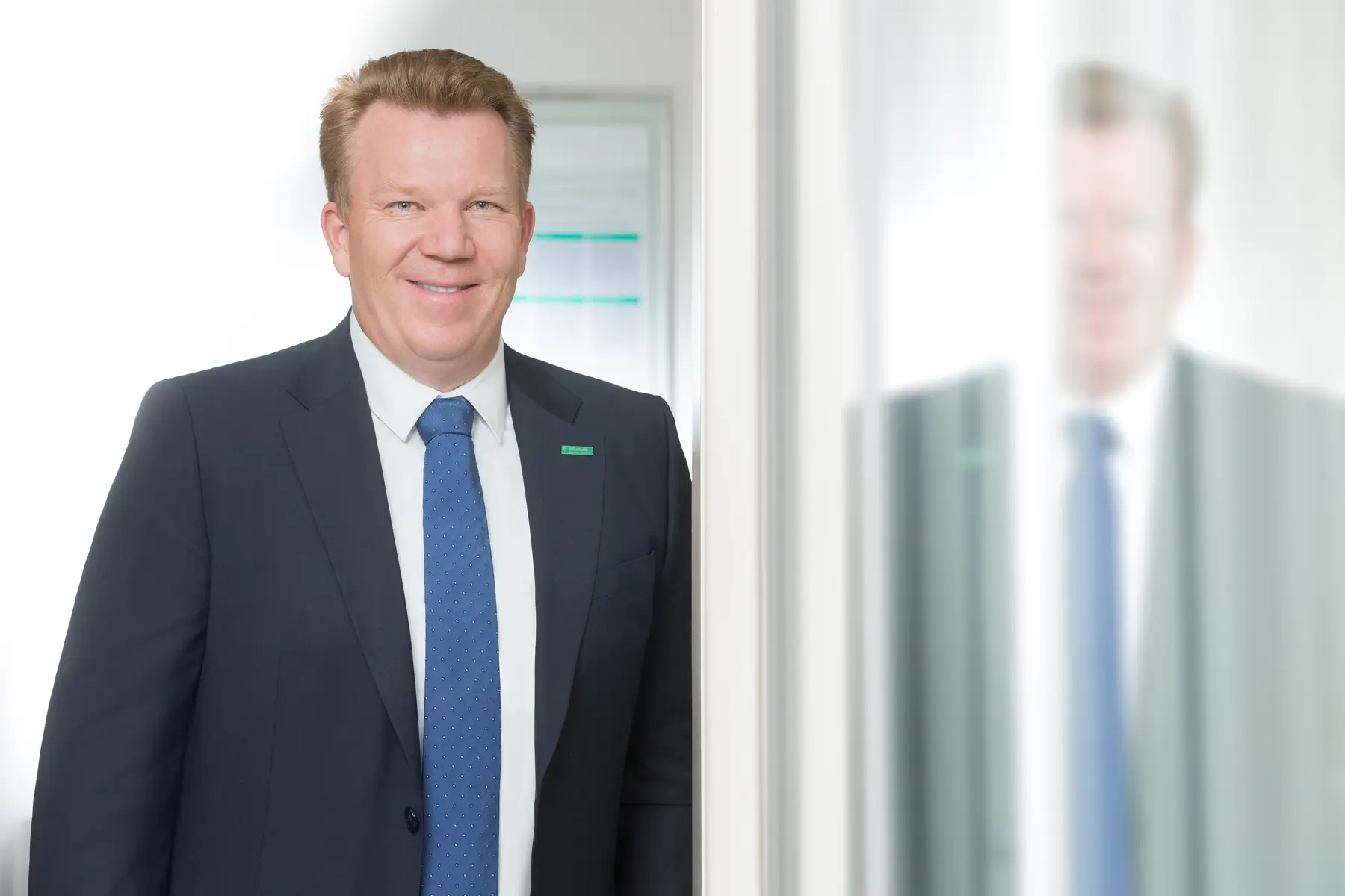 Homem usando terno escuro e gravata azul em ambiente corporativo, posicionado ao lado de uma parede com reflexo.