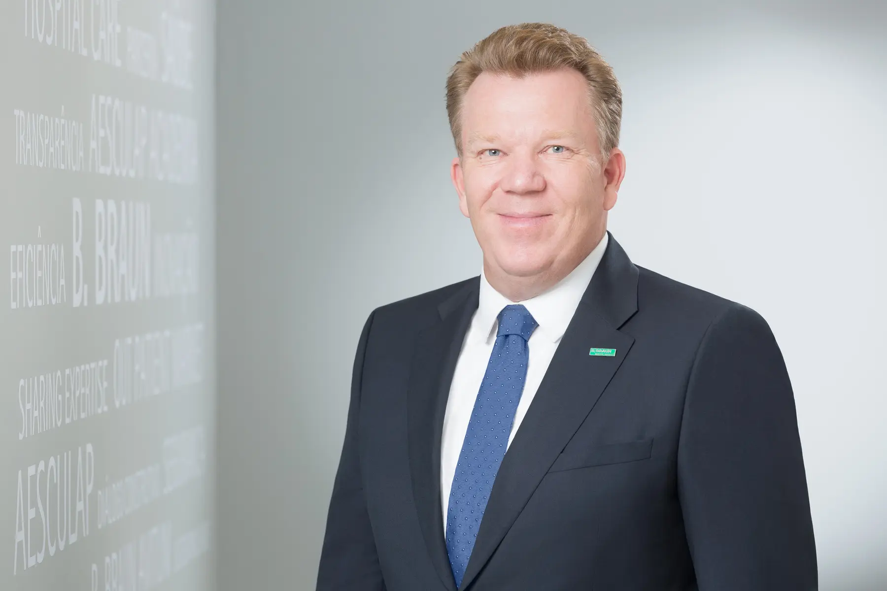 Homem usando terno escuro e gravata azul posicionado diante de parede com palavras institucionais.