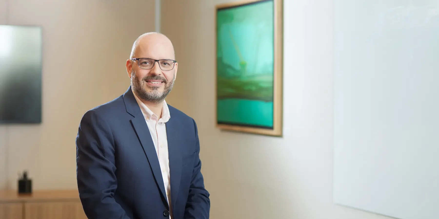 Homem usando blazer azul e camisa clara em sala corporativa com quadro e obra na parede.
