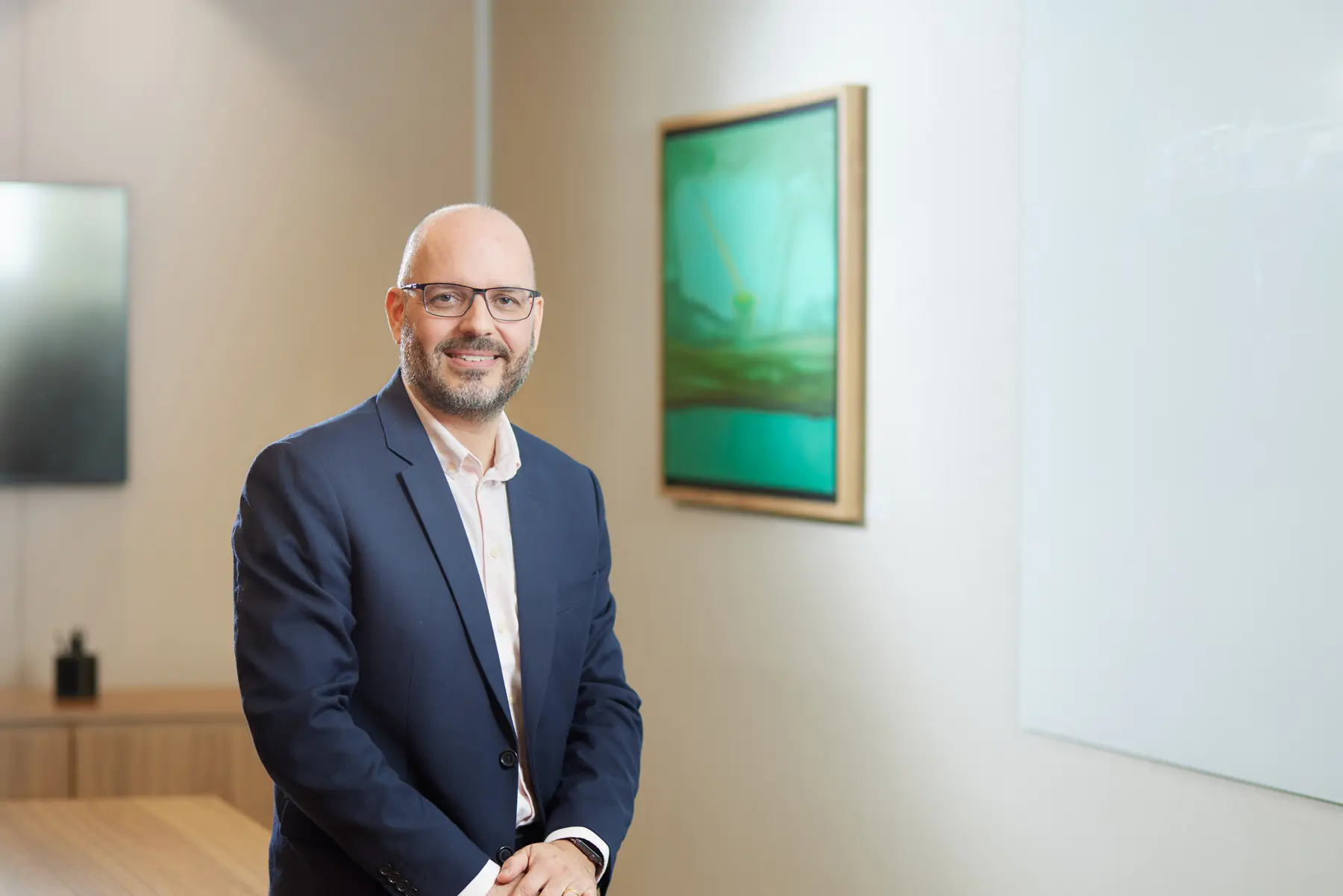 Homem usando blazer azul e camisa clara em sala corporativa com quadro e obra na parede.