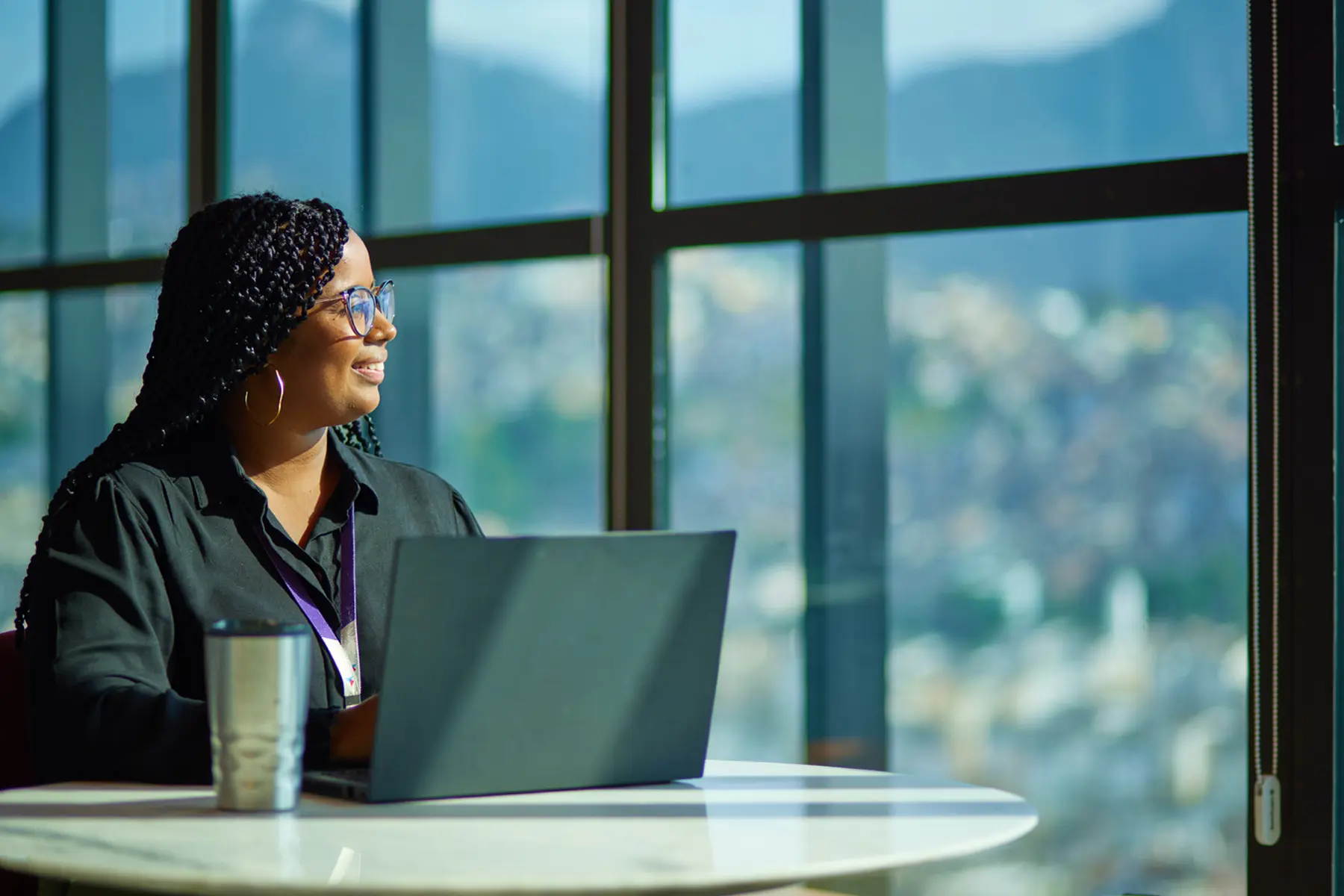 Mulher sentada em frente ao notebook próximo a uma janela ampla com vista desfocada da cidade.