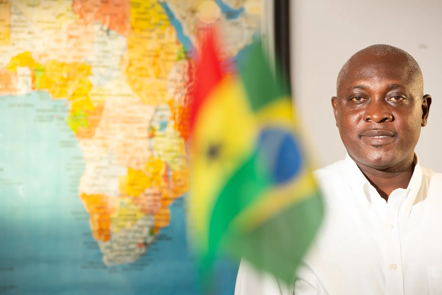 Homem de camisa branca em ambiente interno, em frente a um mapa colorido da África, com bandeira de Gana em primeiro plano.