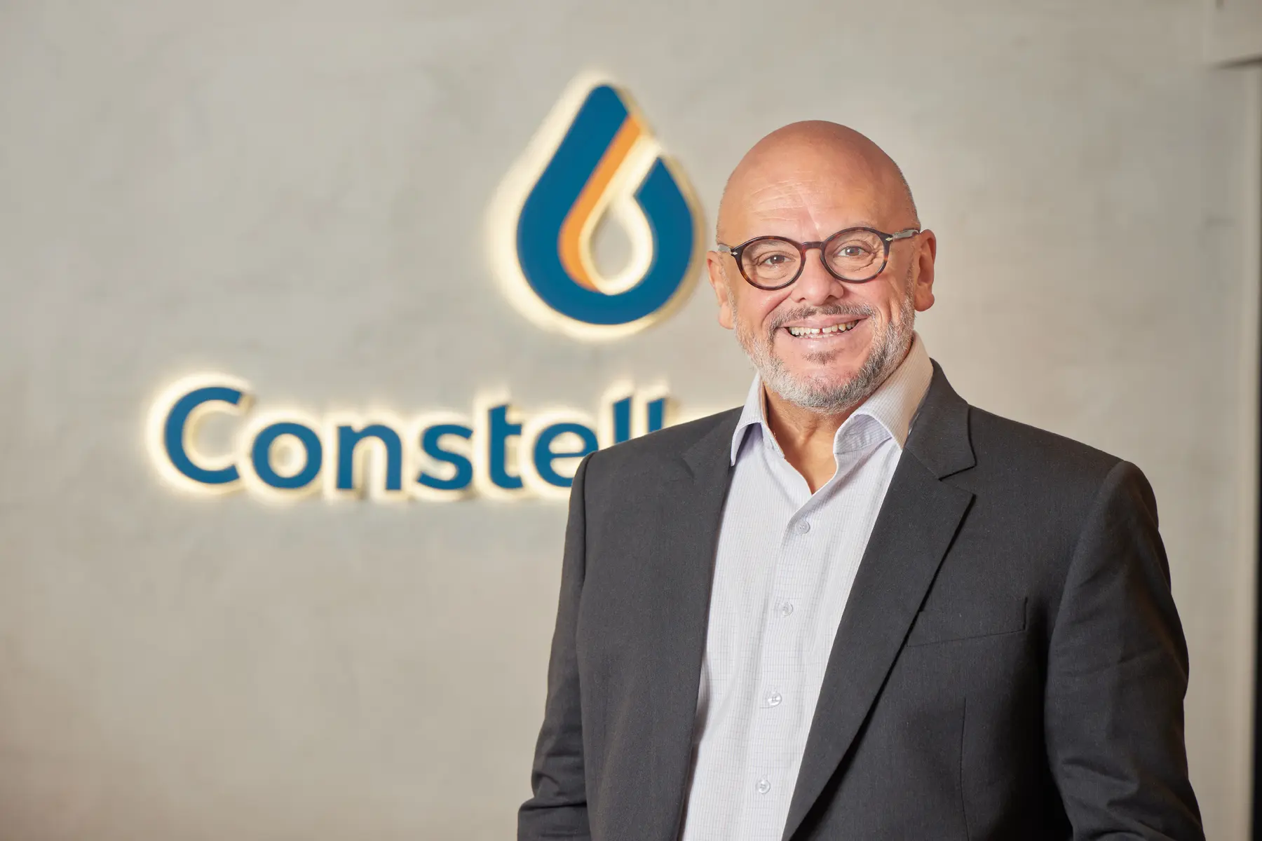 Homem com óculos e blazer escuro em frente a parede com logotipo colorido e texto parcial “Constell”