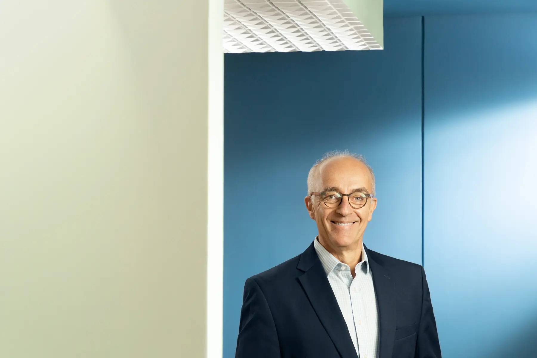 Homem com óculos e blazer escuro em ambiente interno moderno com parede azul e teto branco