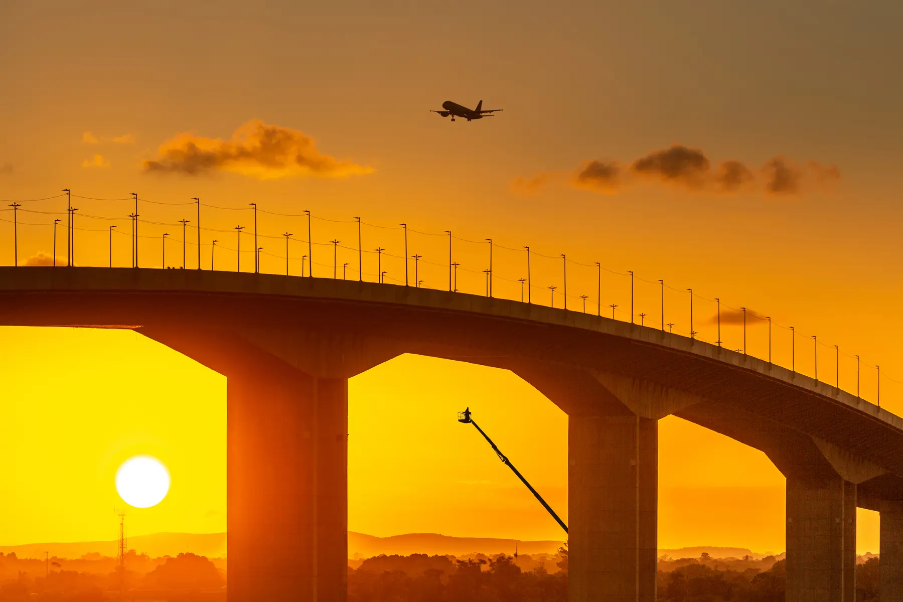 Ponte do Guaíba em Porto Alegre ao pôr do sol, com avião sobrevoando e guindaste abaixo