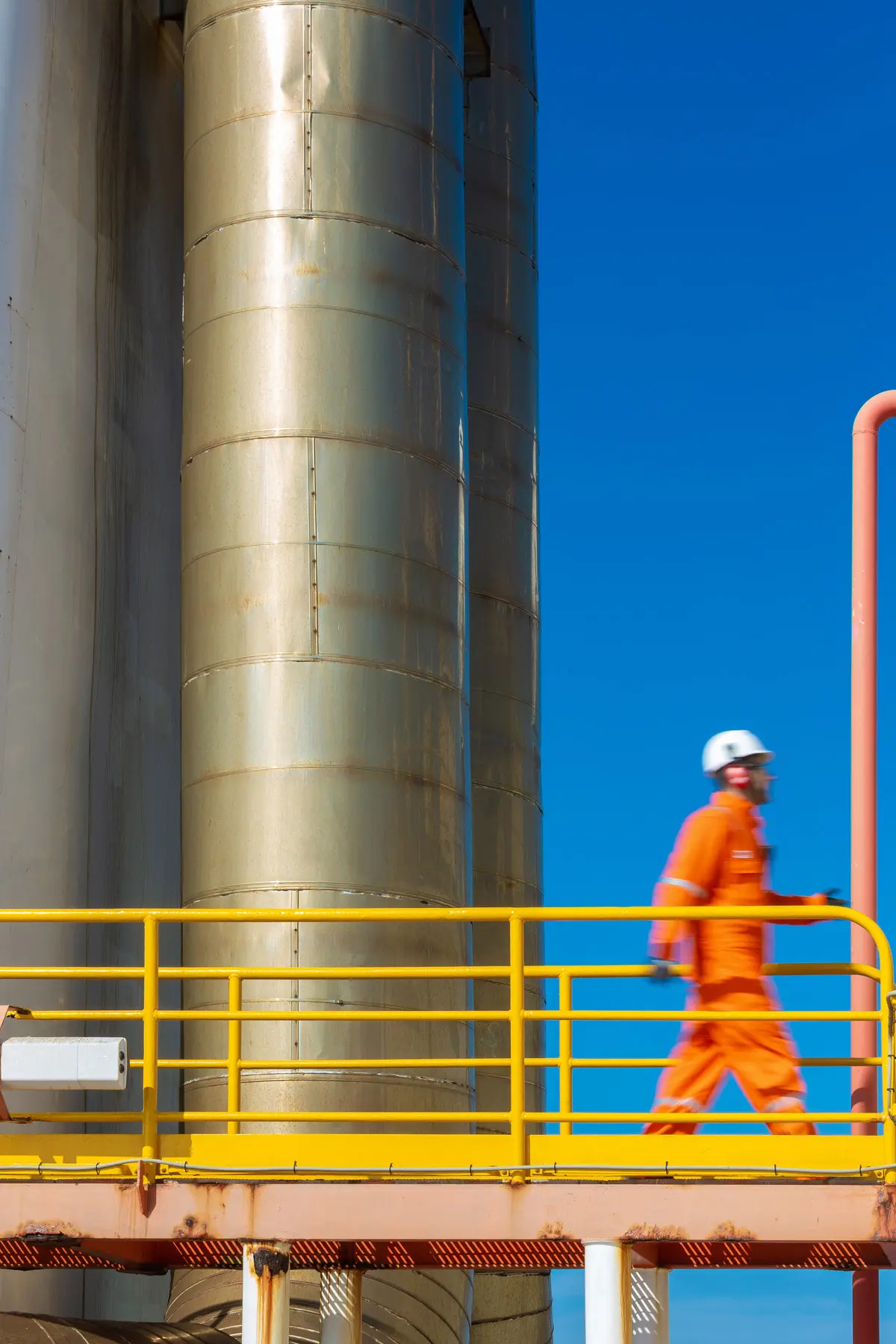 Colaborador caminha sobre passarela metálica em ambiente industrial, utilizando EPI completo sob céu aberto