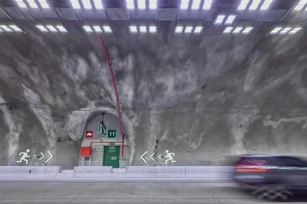 Porta de emergência iluminada em túnel da Rodovia dos Tamoios, com sinalização e veículo em movimento