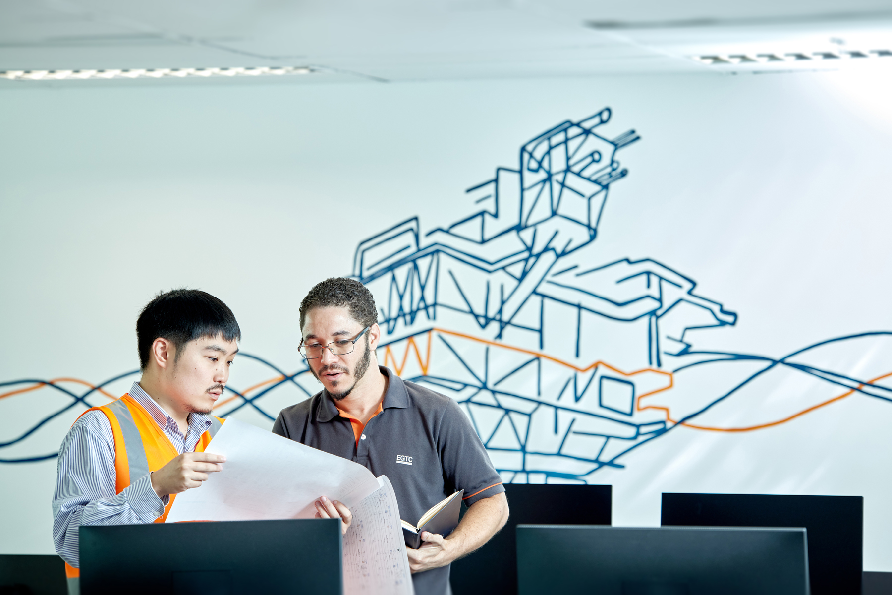 Pessoas com uniforme técnico revisando projetos em escritório com mural de navio e monitores