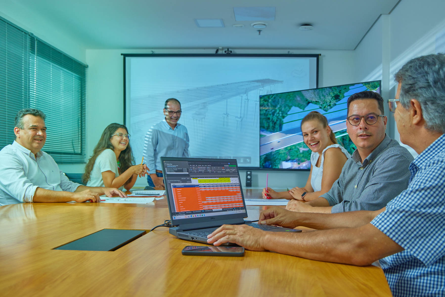 Pessoas reunidas em sala de conferência analisando imagens de infraestrutura e planilha de tarefas
