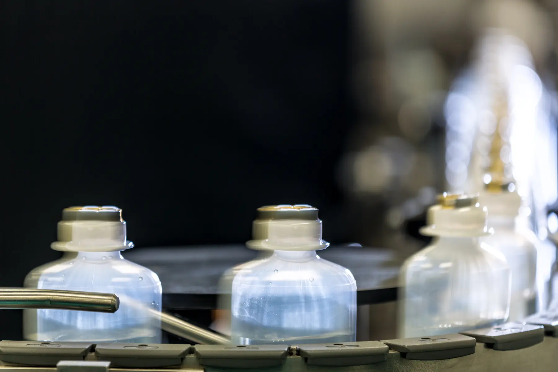 Frascos translúcidos com líquido em movimento sobre esteira automatizada em linha de produção farmacêutica.