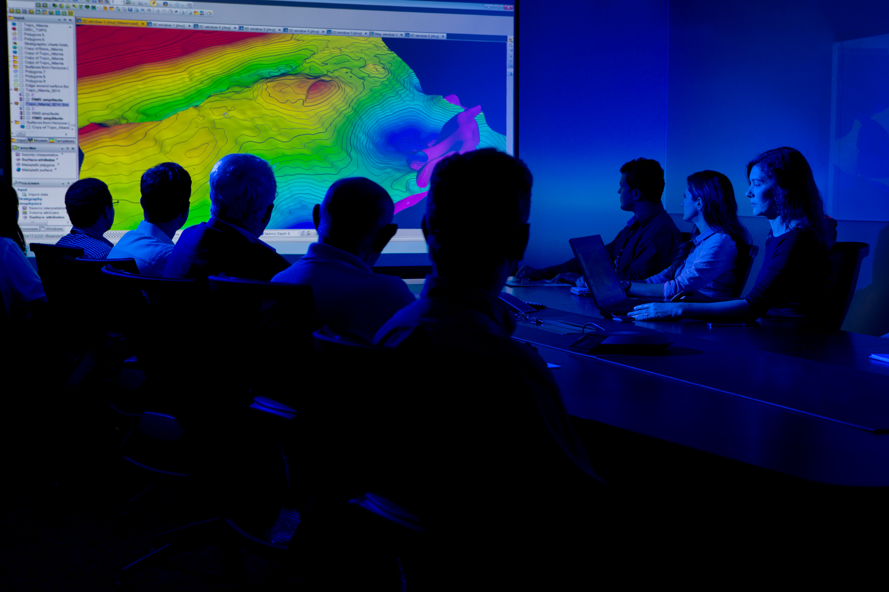 Pessoas em sala escura analisando mapa topográfico digital projetado com dados geoespaciais coloridos