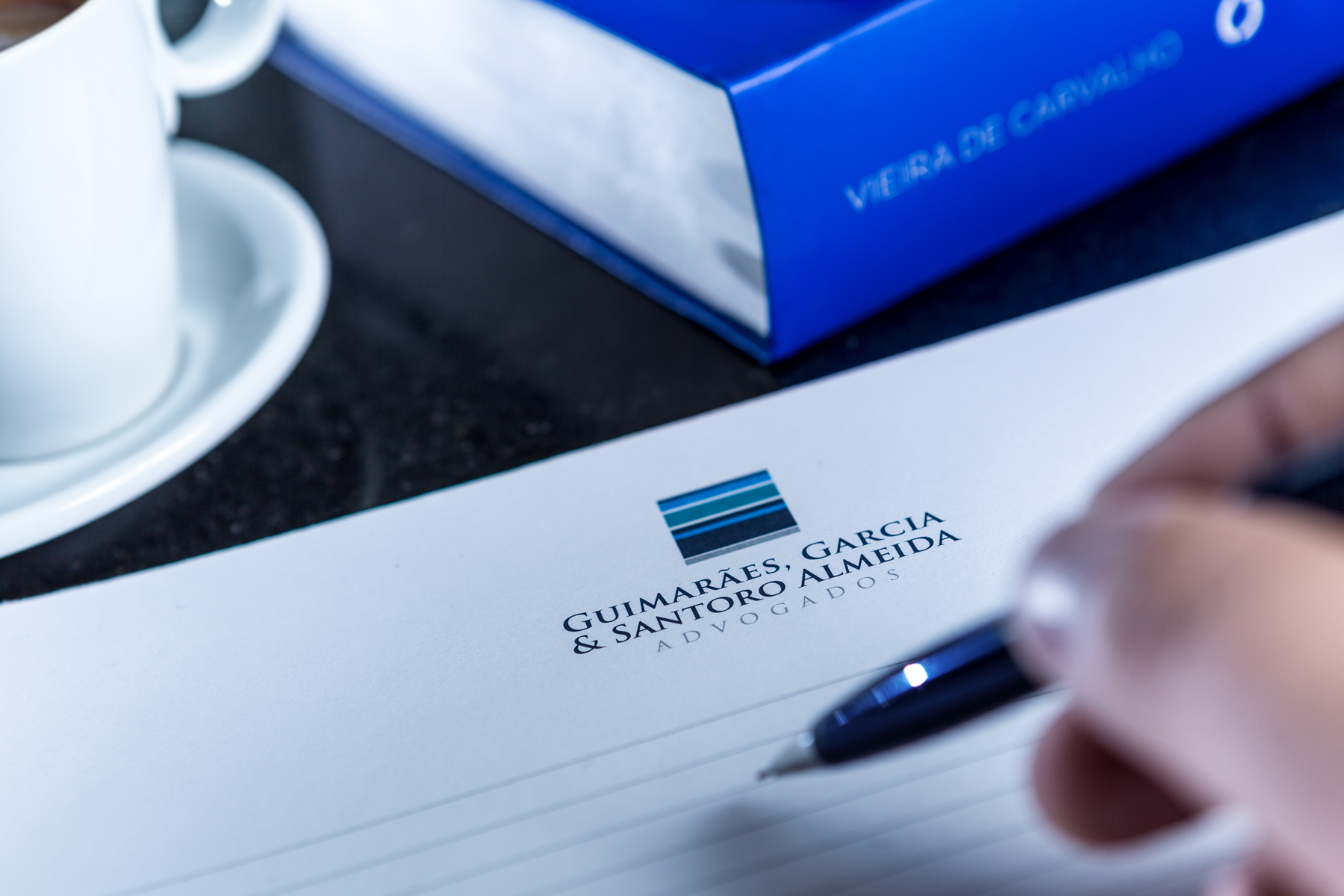 Mão escrevendo em papel timbrado de escritório de advocacia com xícara e livro jurídico ao fundo