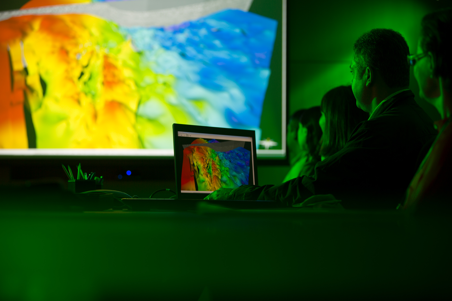 Pessoas analisando visualização 3D de dados geológicos em sala escura com projeção colorida