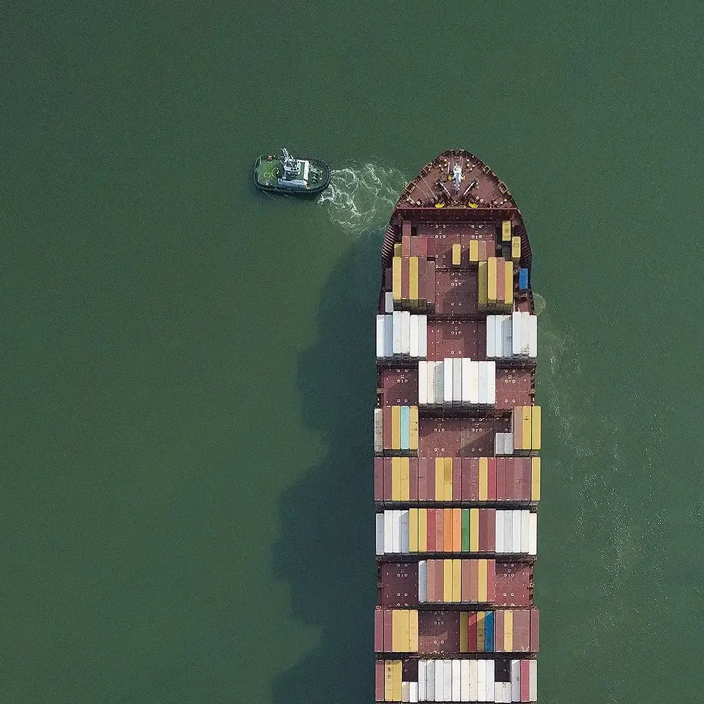 Vista aérea de navio cargueiro, fotografia com drone, com contêineres coloridos sendo acompanhado por rebocador em águas verdes.