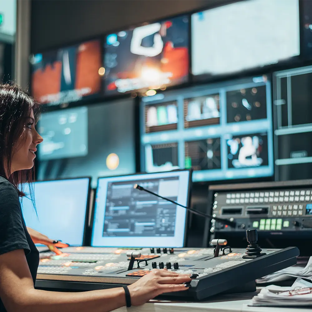 Operador em sala de controle gerenciando painéis e monitores de transmissão de vídeo e áudio.