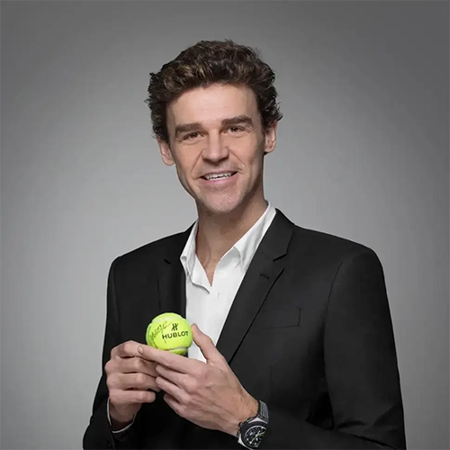 Gustavo Kuerten sorrindo em traje formal, segurando bola de tênis personalizada, em campanha da Hublot
