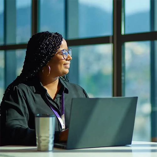 Pessoa com cabelo trançado trabalhando em laptop diante de janela com vista para montanhas e cidade do Rio.