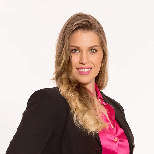 Mulher com blazer preto e blusa pink em retrato profissional sobre fundo branco, com expressão confiante.