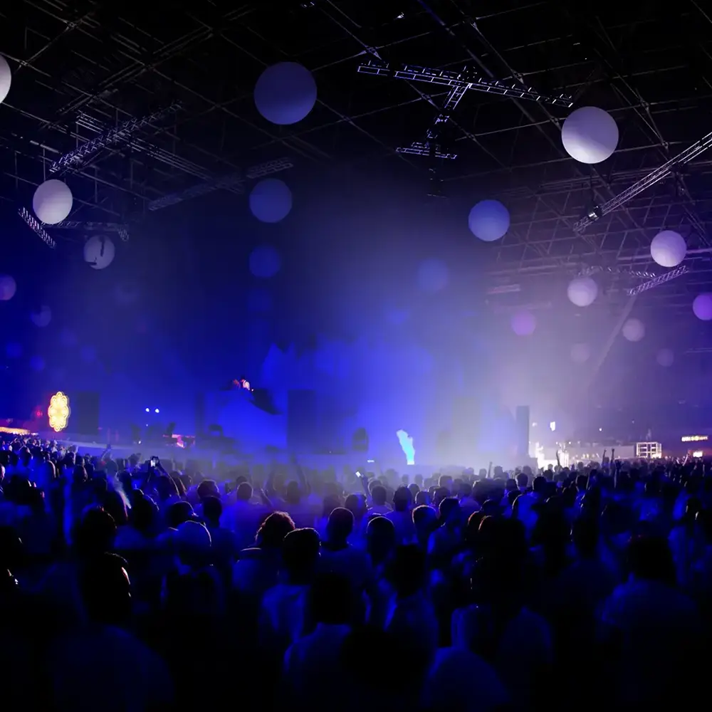 Evento musical com DJ no palco, iluminação azul e púrpura, e público animado em ambiente interno decorado com esferas suspensas.