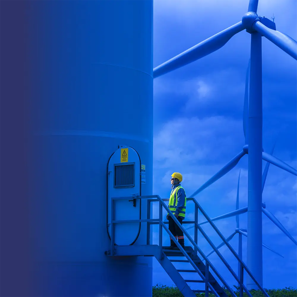 Trabalhador com EPI inspecionando turbina eólica em parque de energia renovável sob céu nublado.