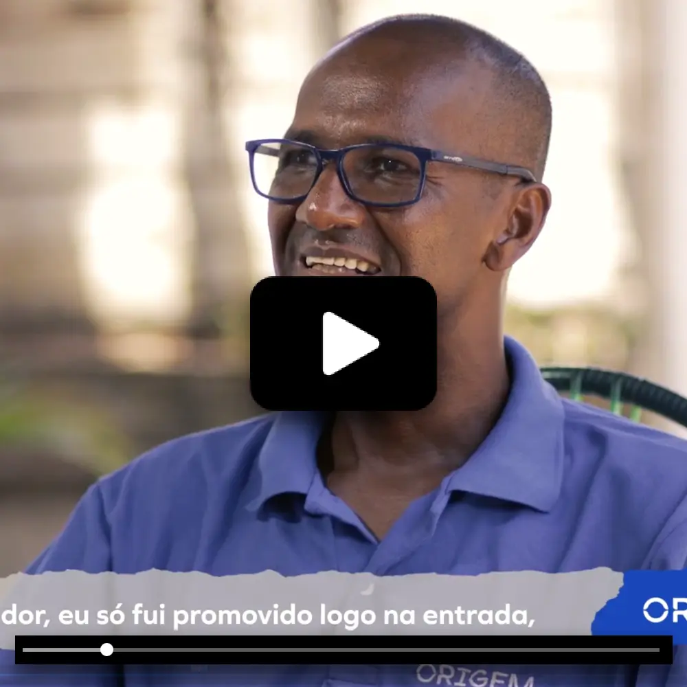 Frame de vídeo com homem de óculos e camisa azul 'ORIGEM', falando em entrevista com legenda em português.