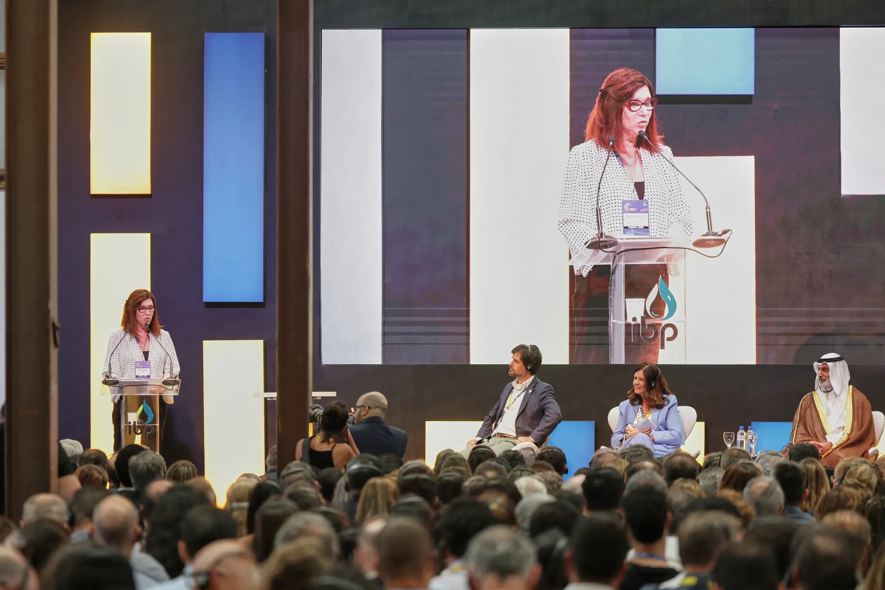 Palestrante feminina, Magda Chambriand, presidente Petrobras, discursa em evento corporativo com público e painel internacional