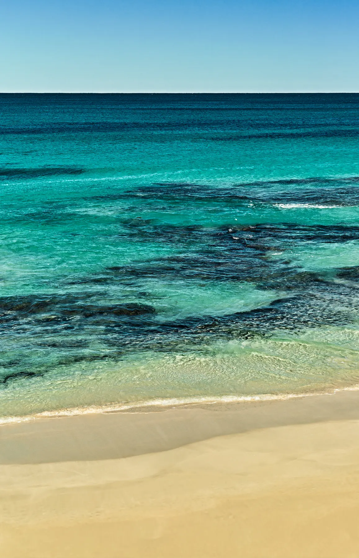 Mar em tons de azul e verde com águas cristalinas tocando a faixa de areia clara da praia